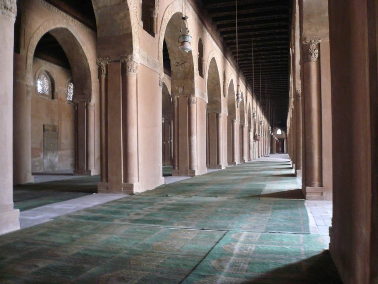A view down a long interior arcade at Ibn Tulun Mosque, Cairo, with rows of pointed arches receding into the distance over a faded green carpet.