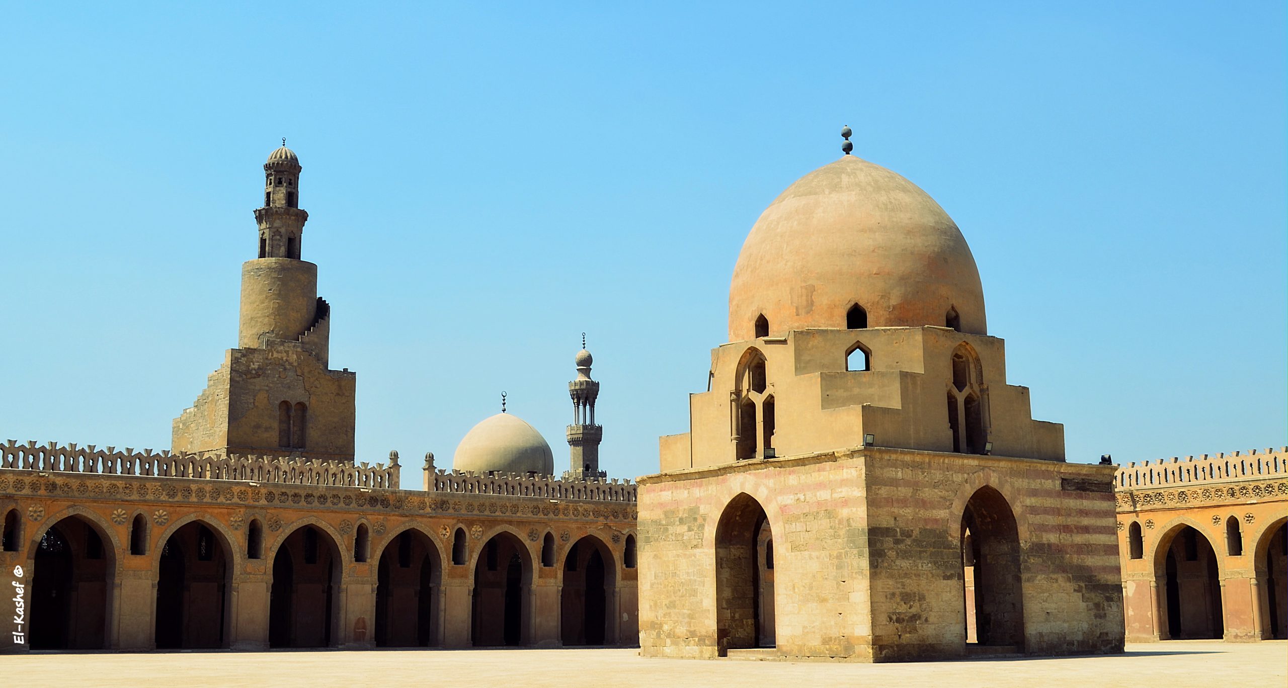 A wide view of the central courtyard of Ibn Tulun Mosque in Cairo, featuring the famous spiral minaret, a secondary minaret in the distance, and the domed structure covering the ancient well in the foreground under a blue sky.