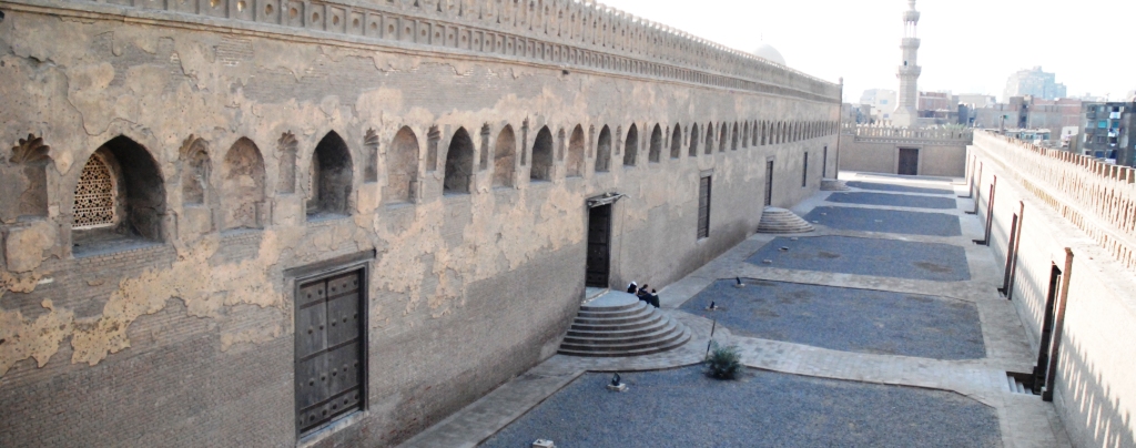 A view of the exterior wall of the Ibn Tulun Mosque from the balcony of the Gayer-Anderson Museum, showing three arched doorways with semicircular steps and three large shuttered windows.