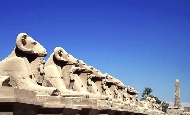 A long, dramatic pathway stretching towards Karnak Temple, flanked on both sides by ancient ram-headed sphinx statues, symbolizing the protection of the god Amun.