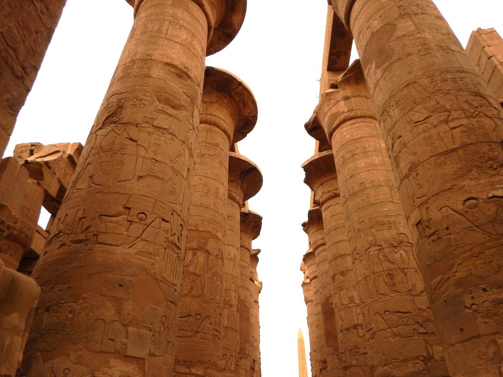 A guest of Mara House looks up at the immense stone columns of the Hypostyle Hall in Karnak Temple, Luxor, as they rise dramatically into a bright, featureless sky