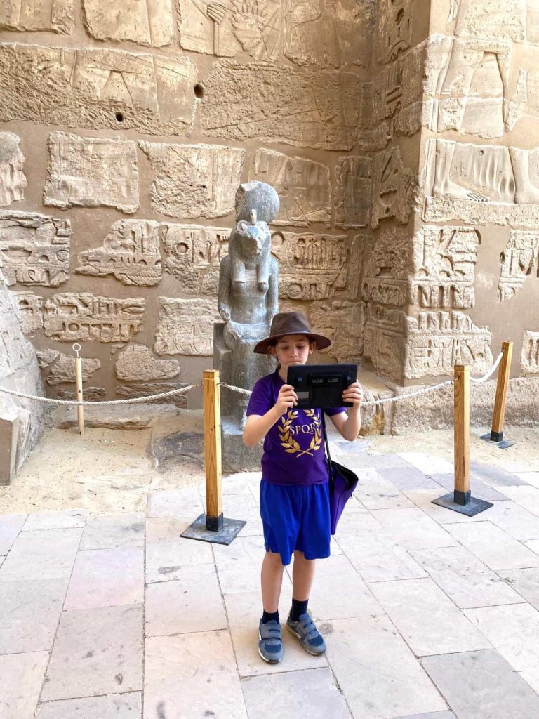 A young boy in an Indiana Jones hat and a purple SPQR t-shirt stands before the massive statue of the goddess Sekhmet at the entrance to Medinet Habu temple in Luxor.