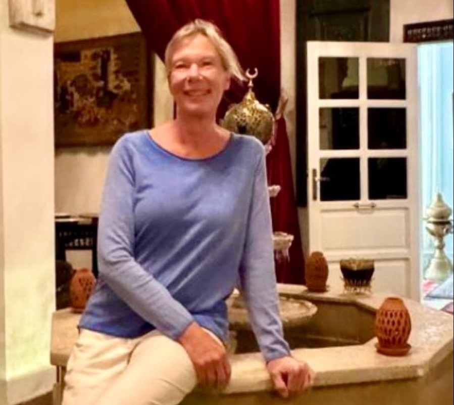 Lady wearing lilac coloured sweater sitting on the edge of a fountain in a large room.