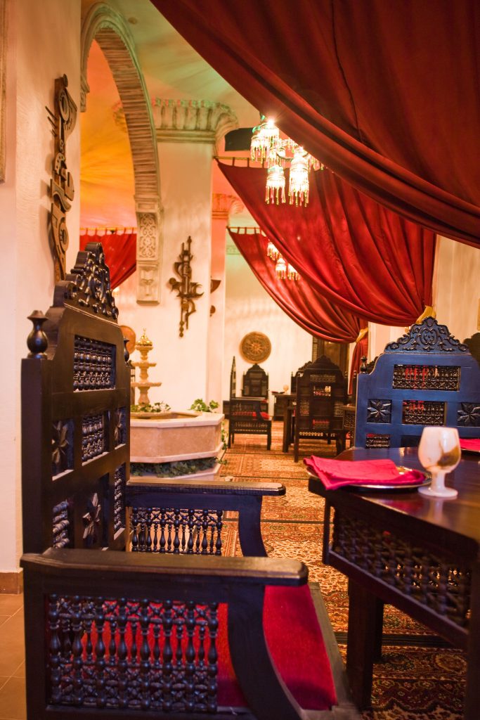 Interior of the Salahadeen Restaurant at Mara House, featuring opulent decor with long red curtains, black wooden tables and chairs, oriental carpets, and a central fountain.