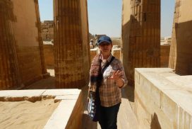 Mara speaking inside the temple complex at Saqqara, sunlight falling across three ancient pillars in the background
