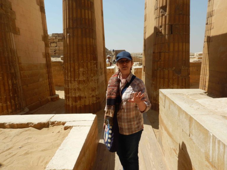 Mara speaking inside the temple complex at Saqqara, sunlight falling across three ancient pillars in the background