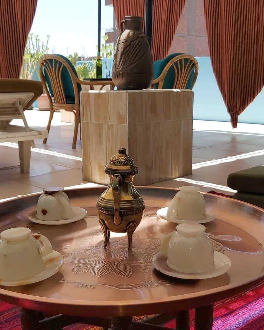 Close-up of a bronze tray with four alabaster coffee cups and a traditional coffee pot on the roof terrace at Mara House Luxor, with tables, plants, chairs, and hanging curtains in the background