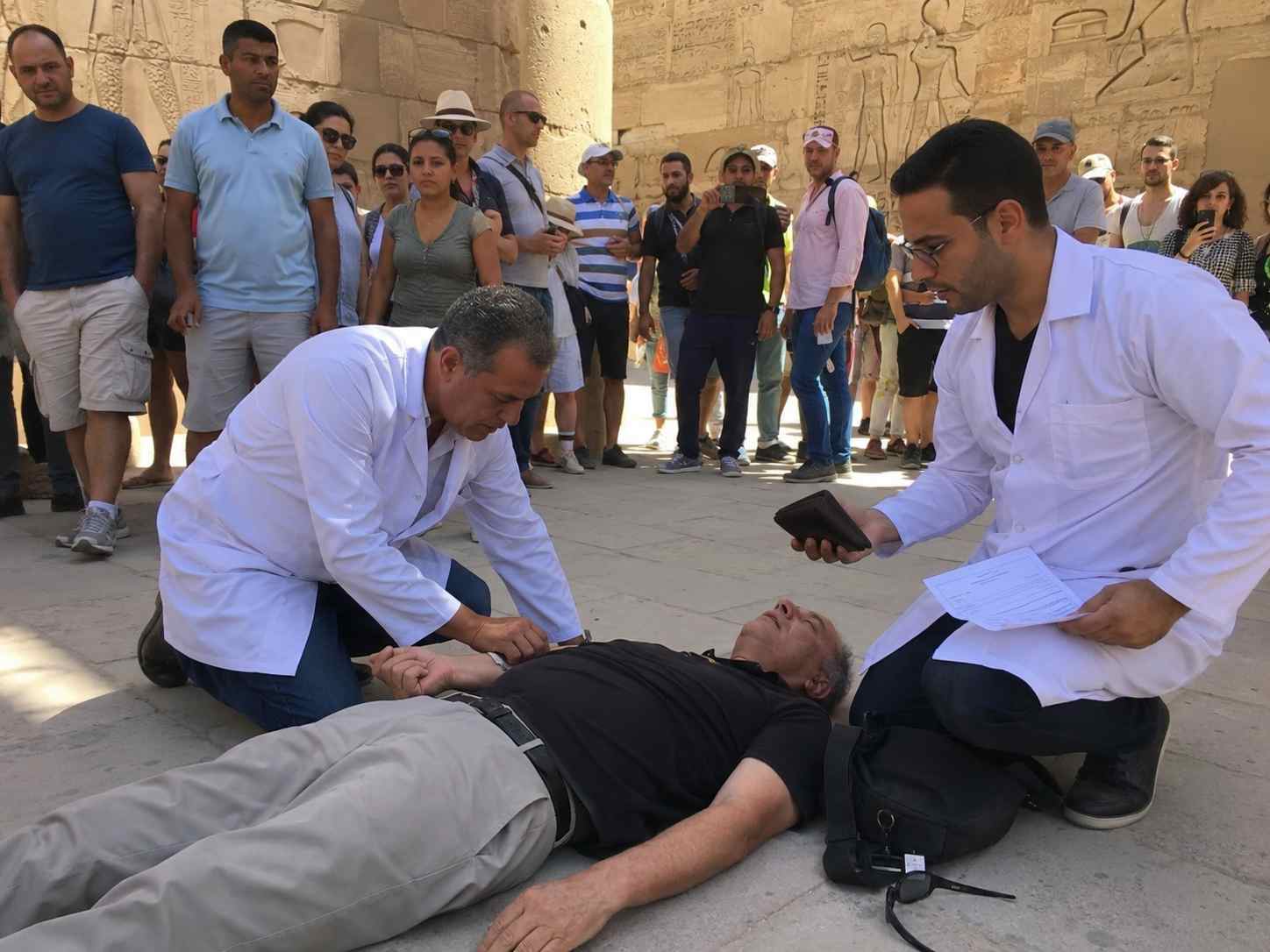 two doctors in white coats kneel beside a man collapsed in from of egyptian temple.