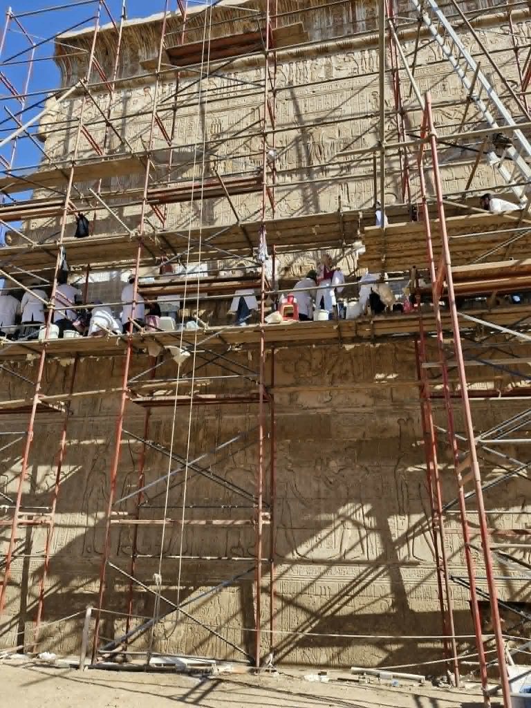 On a high scaffold against the Temple wall a team of white-coated workers are cleaning and restoring the wall
