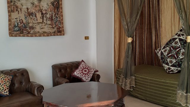 Light & airy living room with extra day bed - romantic atmosphere at Mara House romantic day bed behind long, transparent curtains, green bedcover and large patchwork cushions, 2 brown leather armchairs, sturdy low wooden octagonal table on oriental floor rug