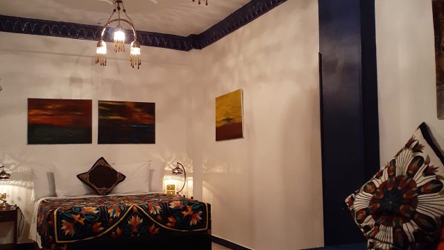 Relax in this calm yet colourful master bedroom at Mara House Bedroom 10m x 4m, double bed covered in handmade patchwork quilt red/black/yelloe/white, bedside lockers with arabian lamps. 4 abstract oil paintings, extra day bed with large patchwork cushions