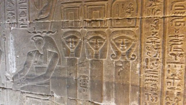 A trinity of becoming. Tucked behind a protective Horus, these three faces of the goddess Hathor are not a repetition, but a progression. Each subtle variation in the styling of her wig suggests a different aspect or stage of her power—perhaps the goddess as maiden, mother, and wise woman; or as the celestial cow of creation, the earthly goddess of joy, and the underworld guide. In a temple of cosmic order, this is a quiet whisper of the goddess's eternal, evolving nature. Close-up carving in the Dendera crypt of three Hathor heads, each with a slightly different hairstyle, arranged behind a seated figure of the god Horus.